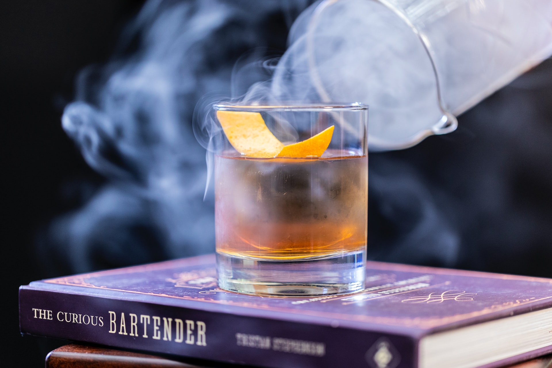 Old-fashioned cocktail in glass placed on book "The Curious Bartender" with smoke being poured on top