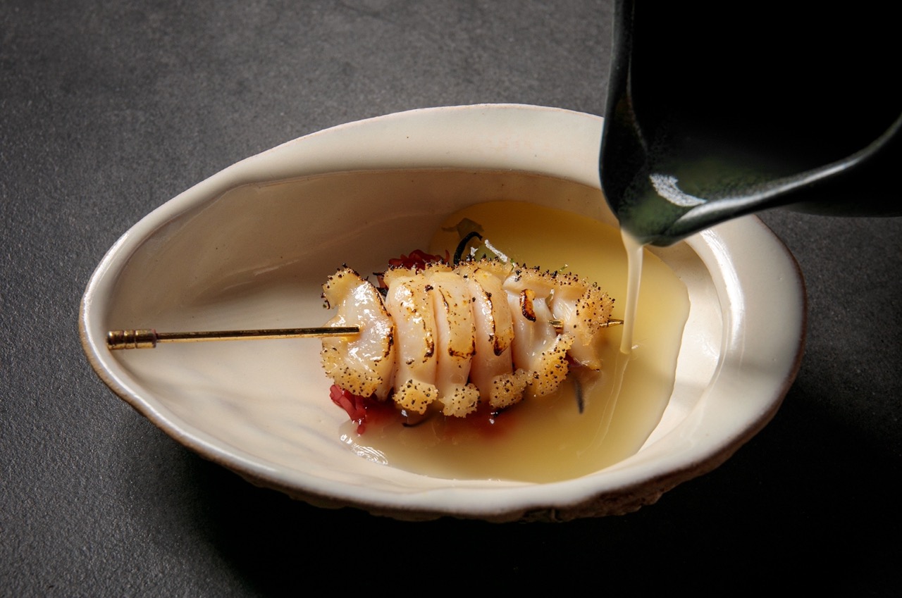 A green jug pouring sauce over an abalone skewer in an irregularly-shaped white bowl