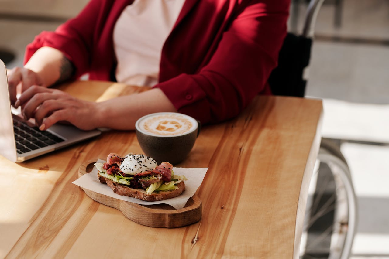 Wheelchair user using a laptop with a coffee and poached egg on toast beside her