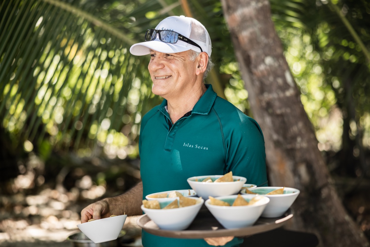 A smiling server wearing a uniform of a t-shirt and baseball cap, carrying a tray of bowls in the sunshine with palm trees in the background