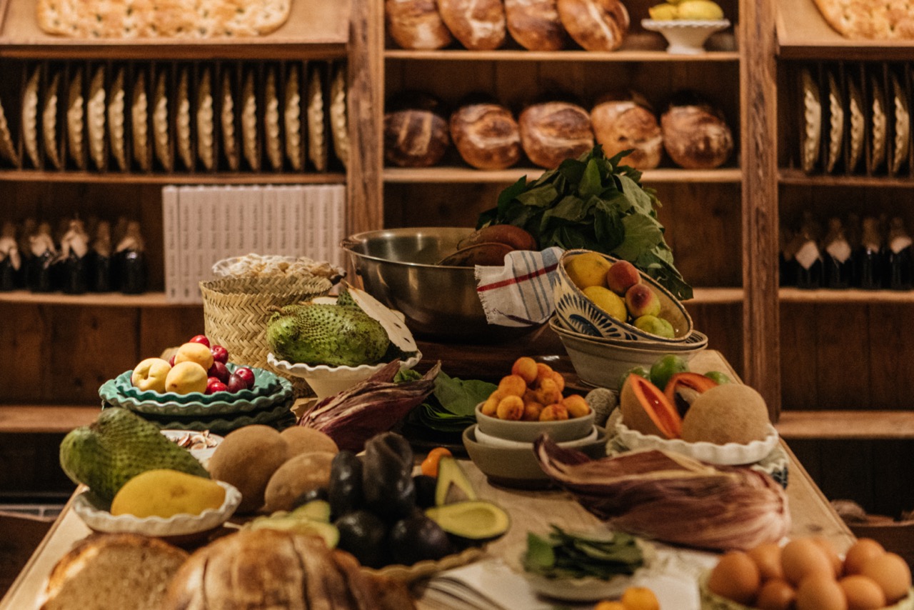 Table heaped with fresh Mexican produce, with racks of bread loaves in the background