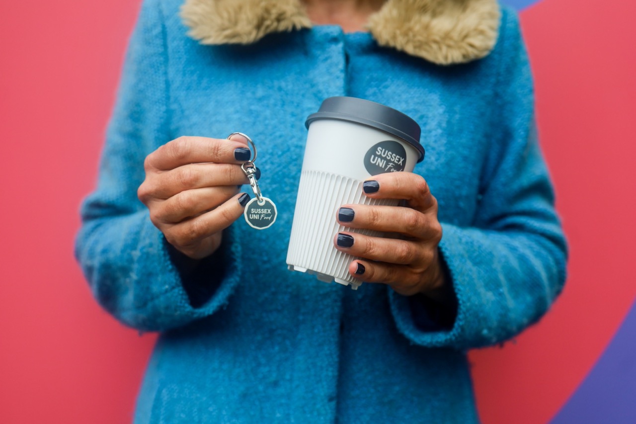 Person in blue cardigan holding University of Sussex keychain and resuable cup against pink background