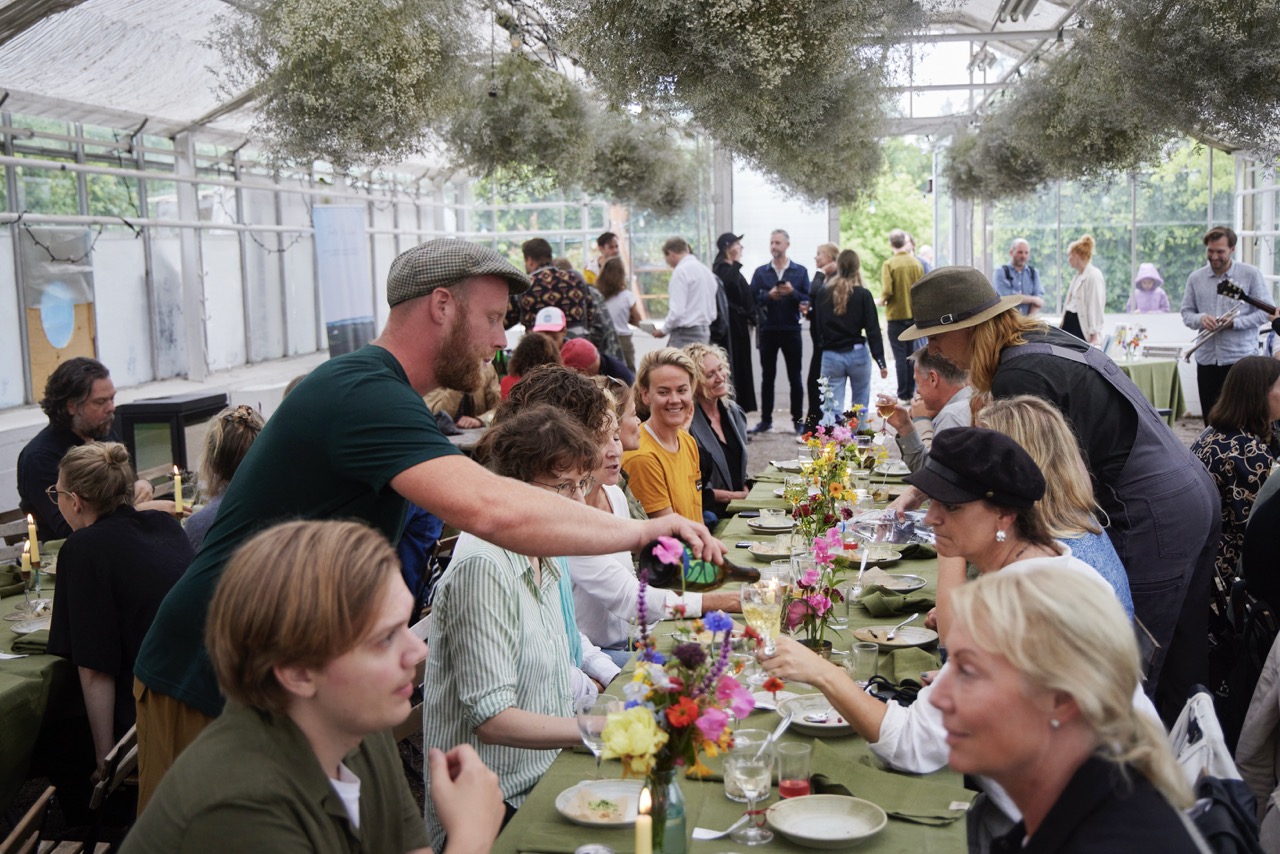 A table full of people enjoying a meal together in a bright room with a glass roof and hanging plants