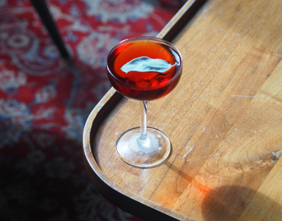 An elegant red-coloured cocktail in a stemmed glass on the corner of a wooden table, with patterned carpet in the background