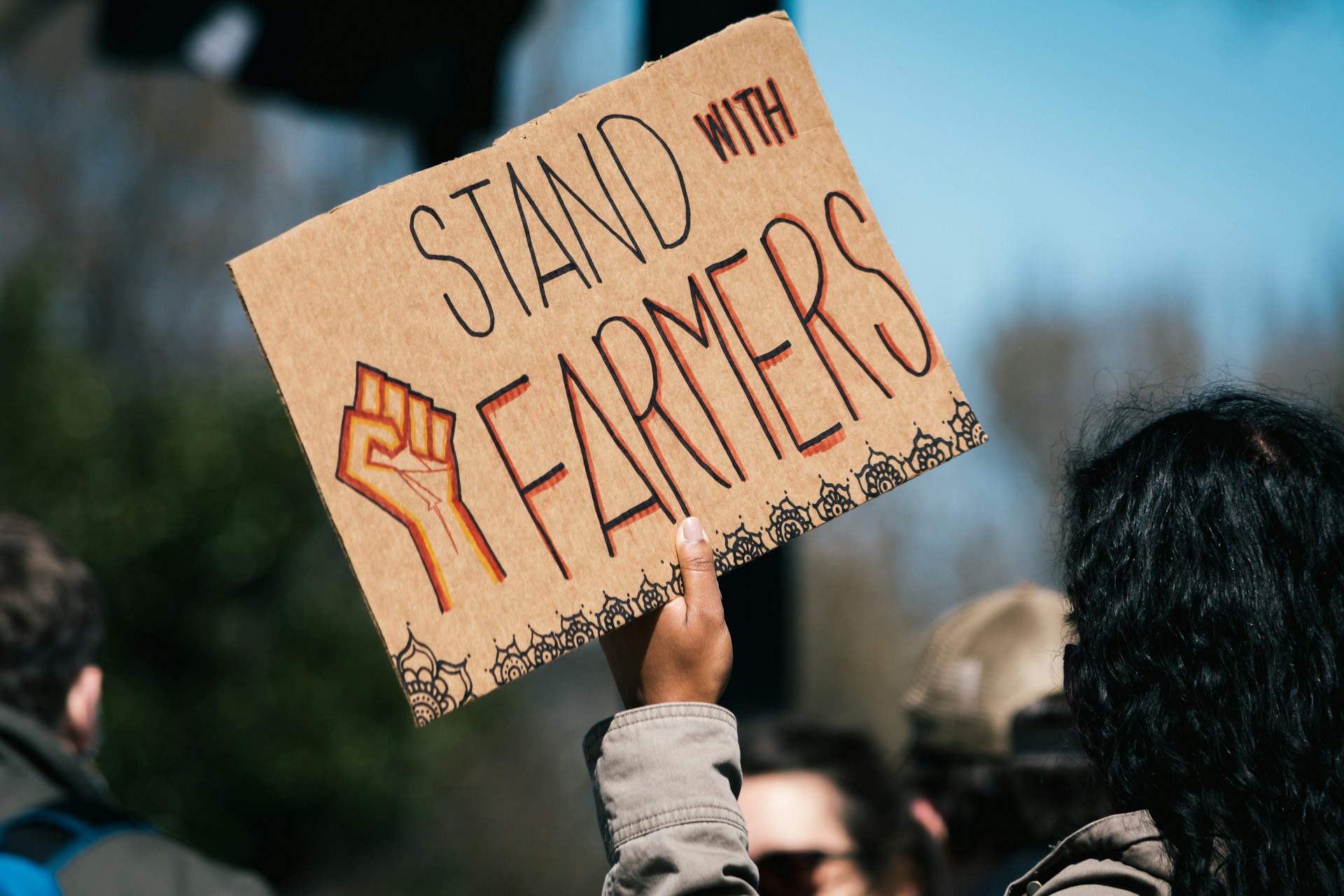 A person with long black hair holding up a sign that says 'stand with farmers' with a drawing of a clenched fist