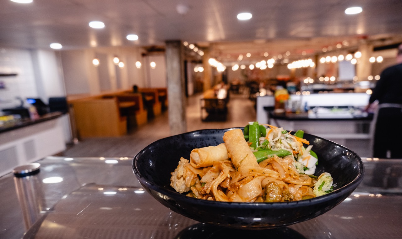 Bowl of vegan pad see ew with tofu, mangetout and spring rolls, with college canteen in background