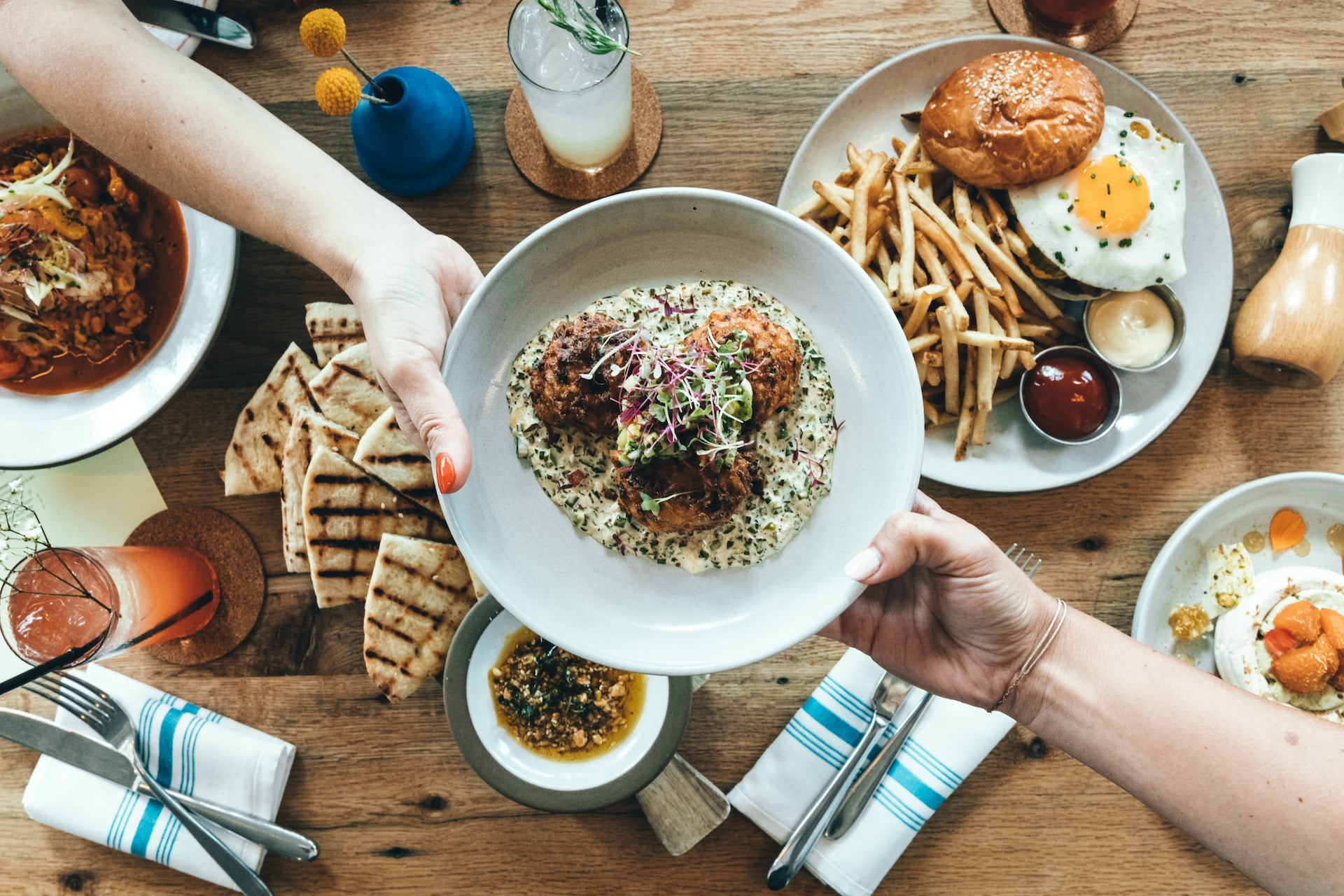 Hands passing a bowl of food across a table. The table has other plates of food and drinks on it, including a burger with a fried egg and chips