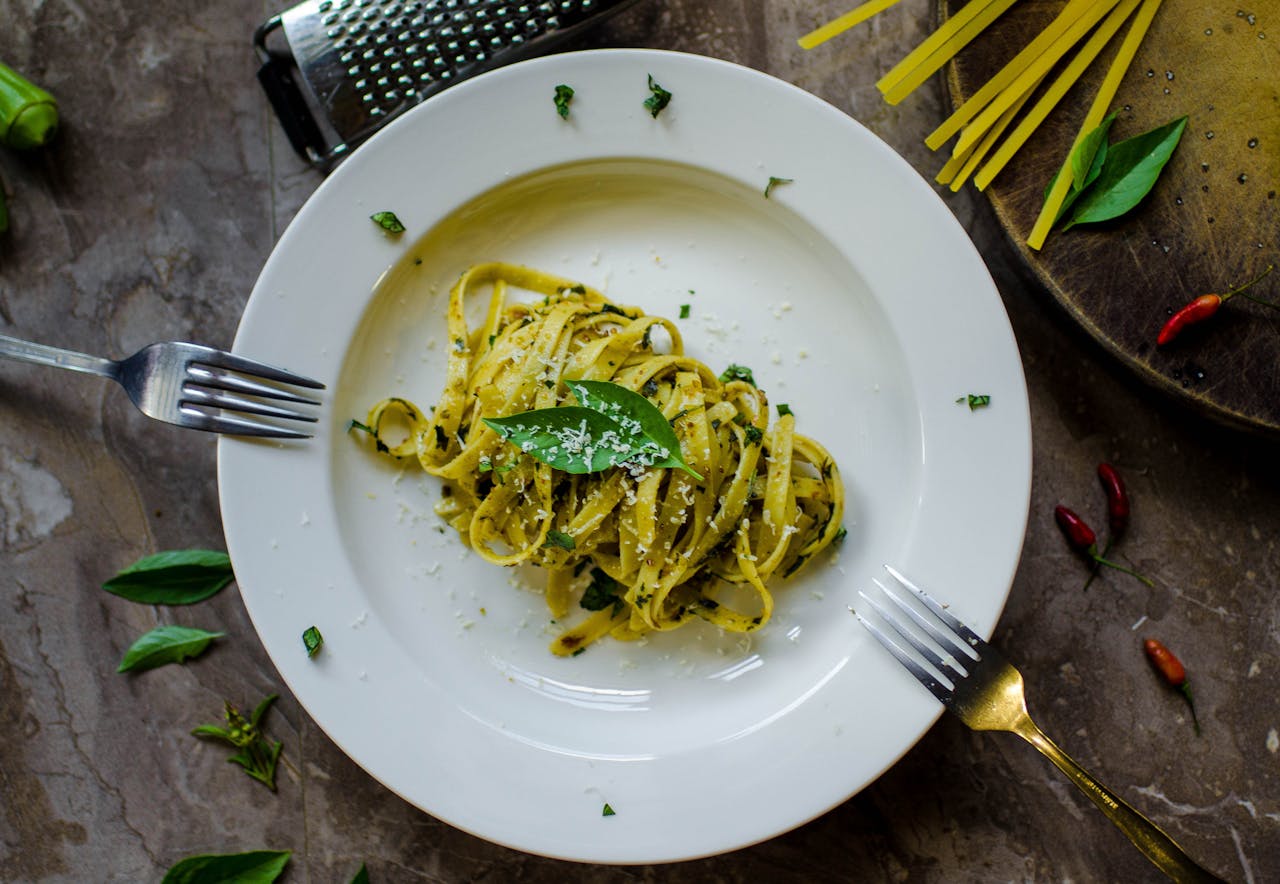 Overhead shot of vegetarian pasta dish with basil leaves, with two forks resting against plate edge and cheese grater beside it