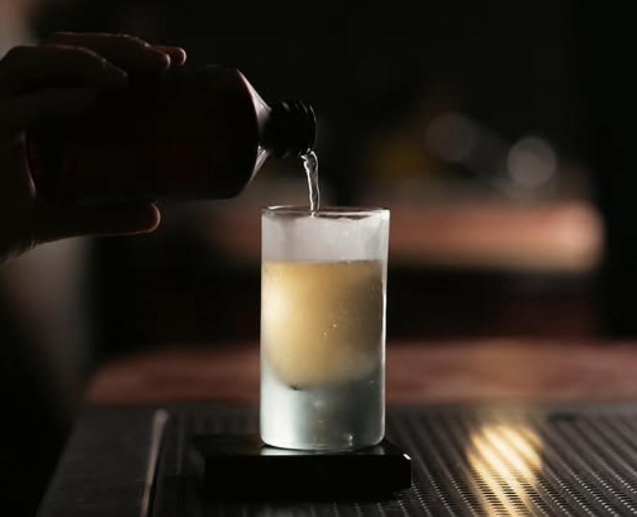 A person pouring a pale yellow cocktail into a chilled tumbler in a darkened room