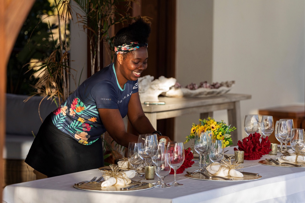 A smiling server in a uniform with colourful flowers, setting a table in the sunshine