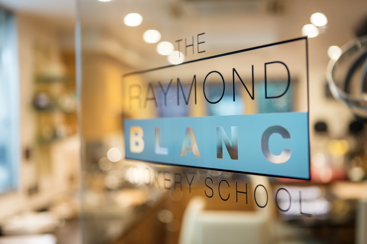 Raymond Blanc Cookery School sign on glass door, with kitchen blurred in background