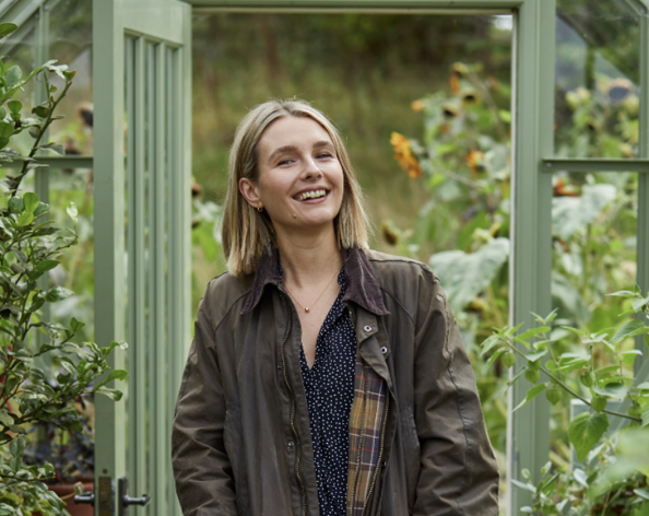 Kate Harvey against a background of foliage with orange flowers and a greenhouse doorway