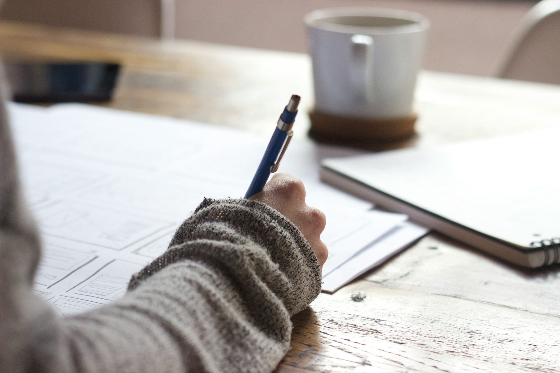 A woman's hand holding a pen and writing, with lots of papers on the desk and a cup in the background