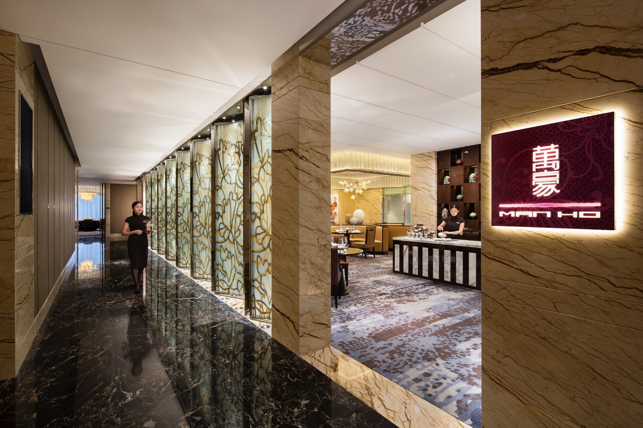 A hotel corridor with a member of staff walking in the background and a sign in the foreground saying 'MAN HO'
