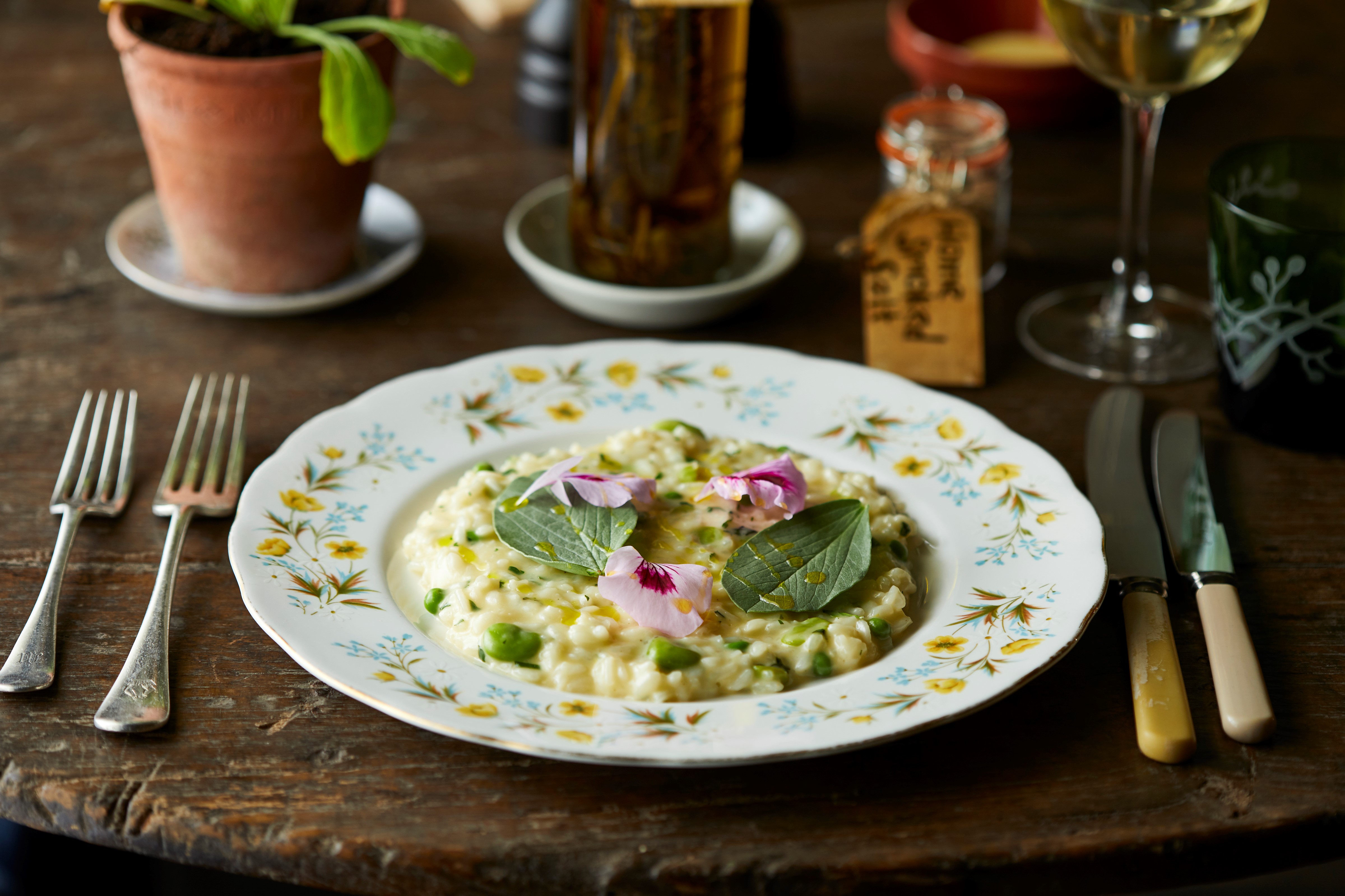 Pea risotto garnished with flowers on a table with cutlery and a glass of wine.
