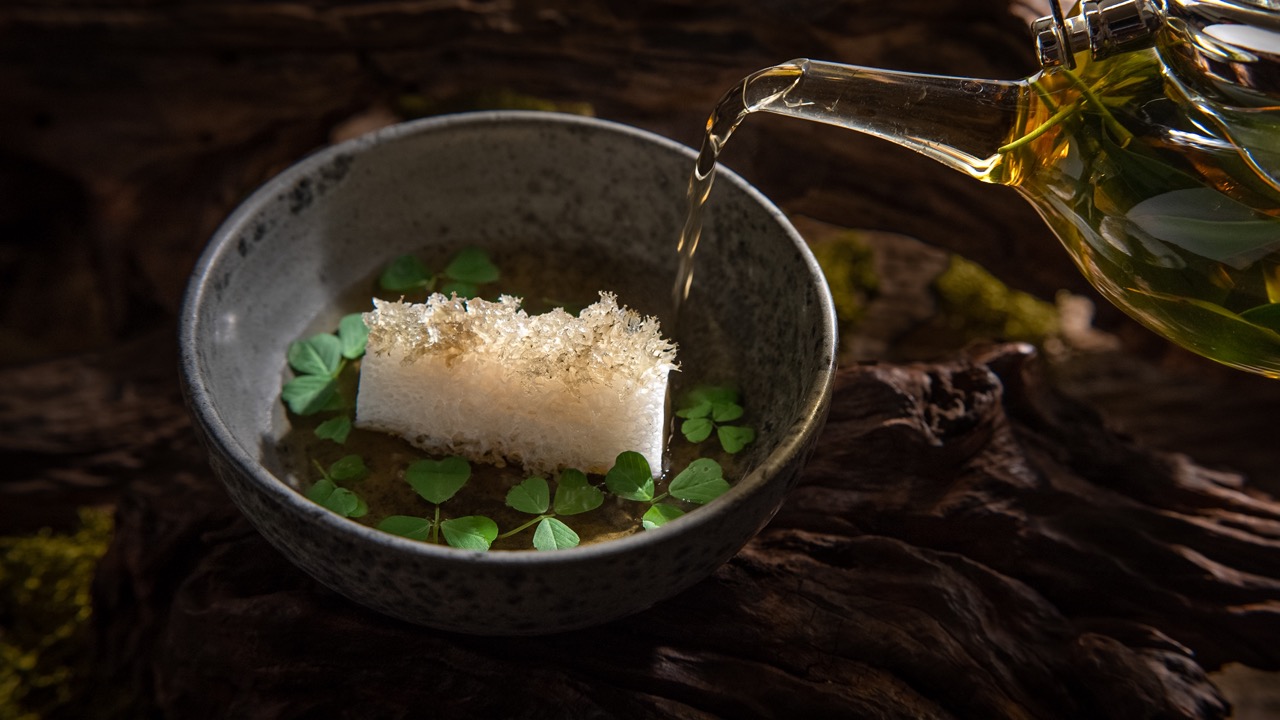 Golden liquid being poured from a glass jug into a bowl of beautiful food against a dark background