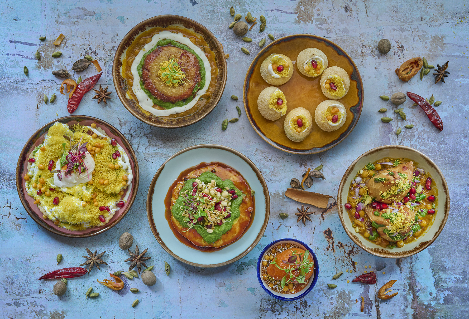 Overhead shot of Indian food in bowls against bright blue table, with chillies and star anise scattered around