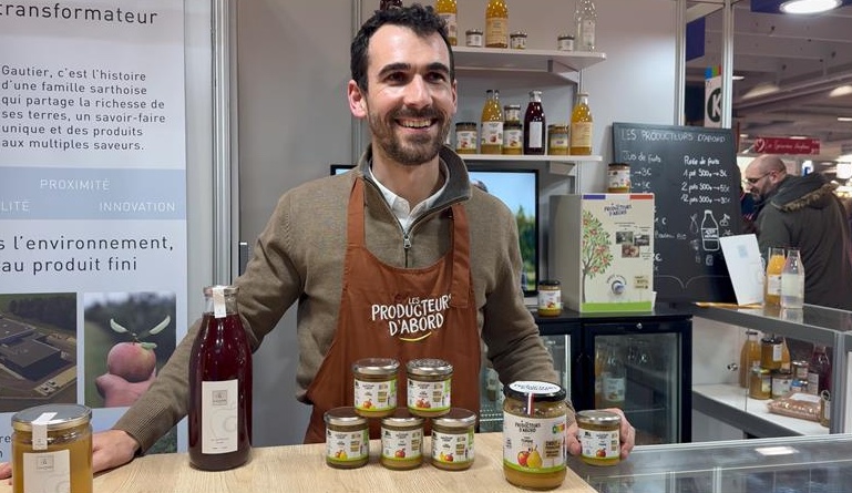 A smiling man wearing a "Producteur d'Abord" apron, with pots of preserves