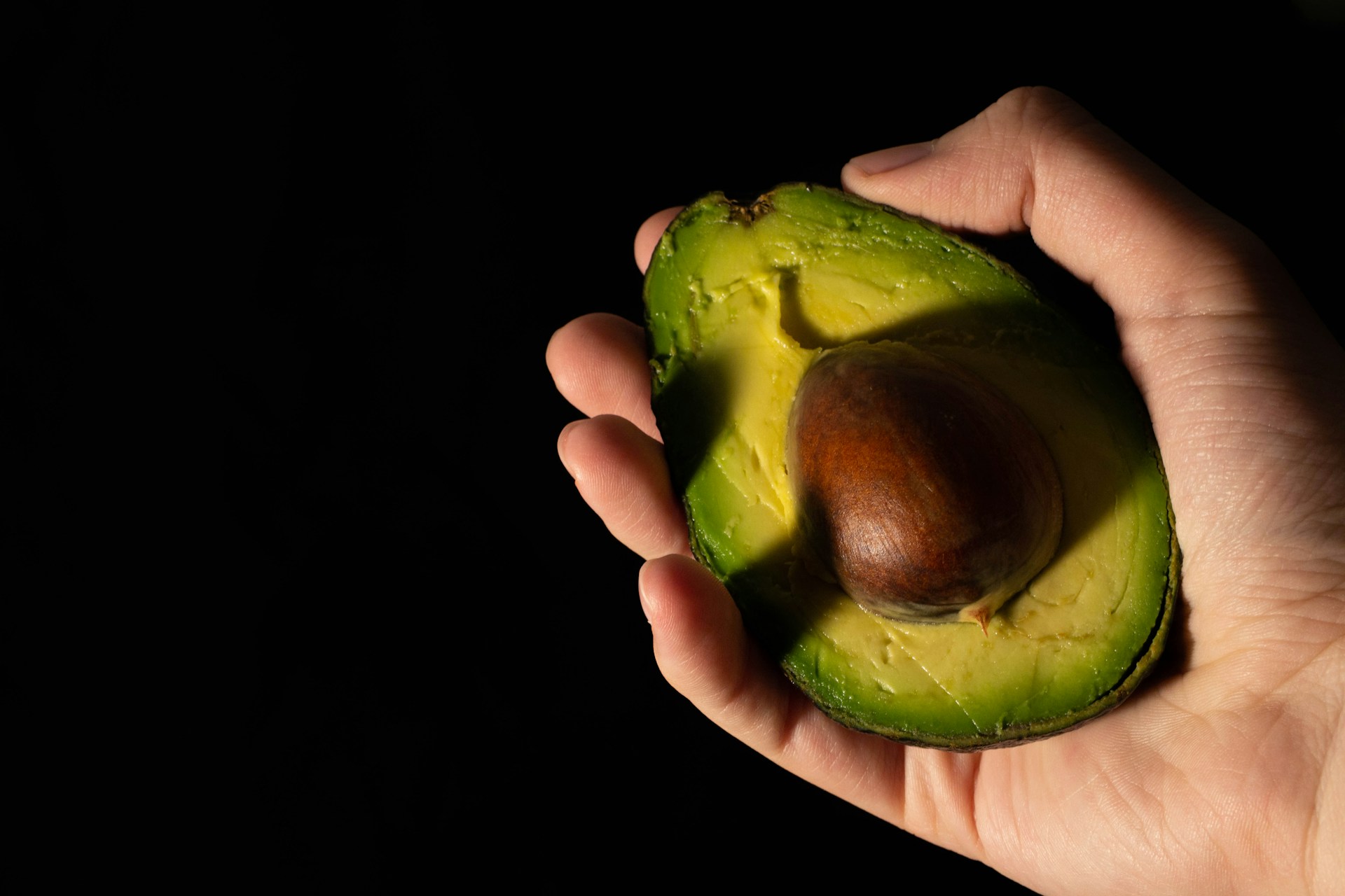 A hand holding half an avocado against a dark background
