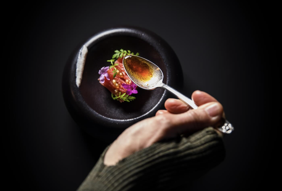Overhead shot of a bowl of beautiful, colourful food against a very dark background, with a hand holding a spoon