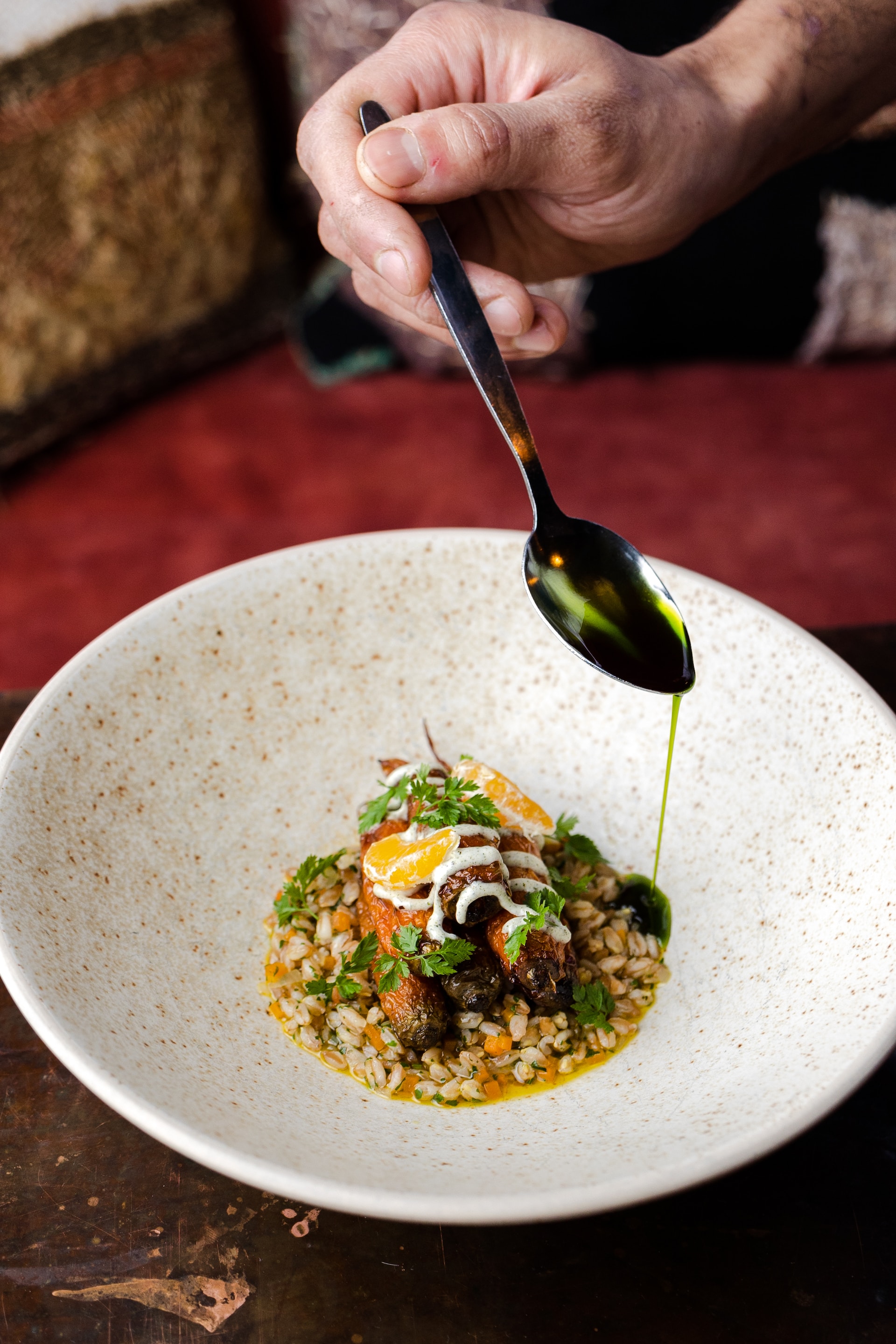 Chef's hand drizzling herb oil from spoon over barley and sausage dish