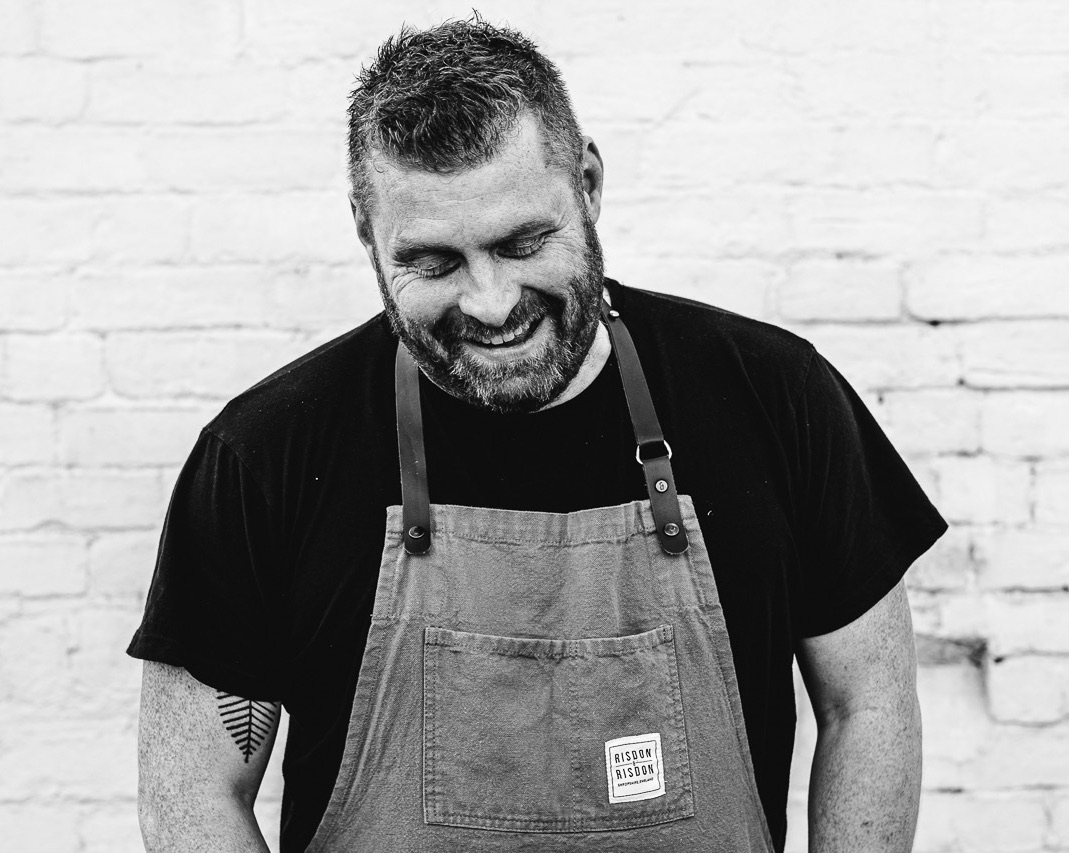 Black and white image of Chef James Sherwin of Wild Shropshire, laughing in front of brick wall