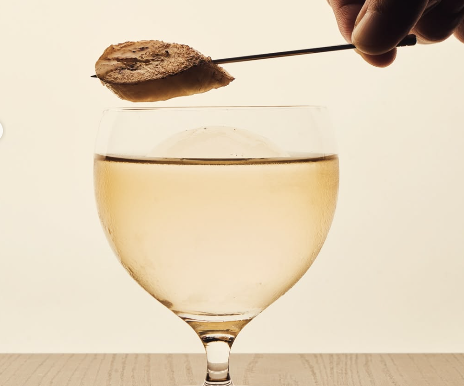 A hand holding a biscuit on a cocktail stick over a pale golden drink in a glass, at Bar Us in Bangkok
