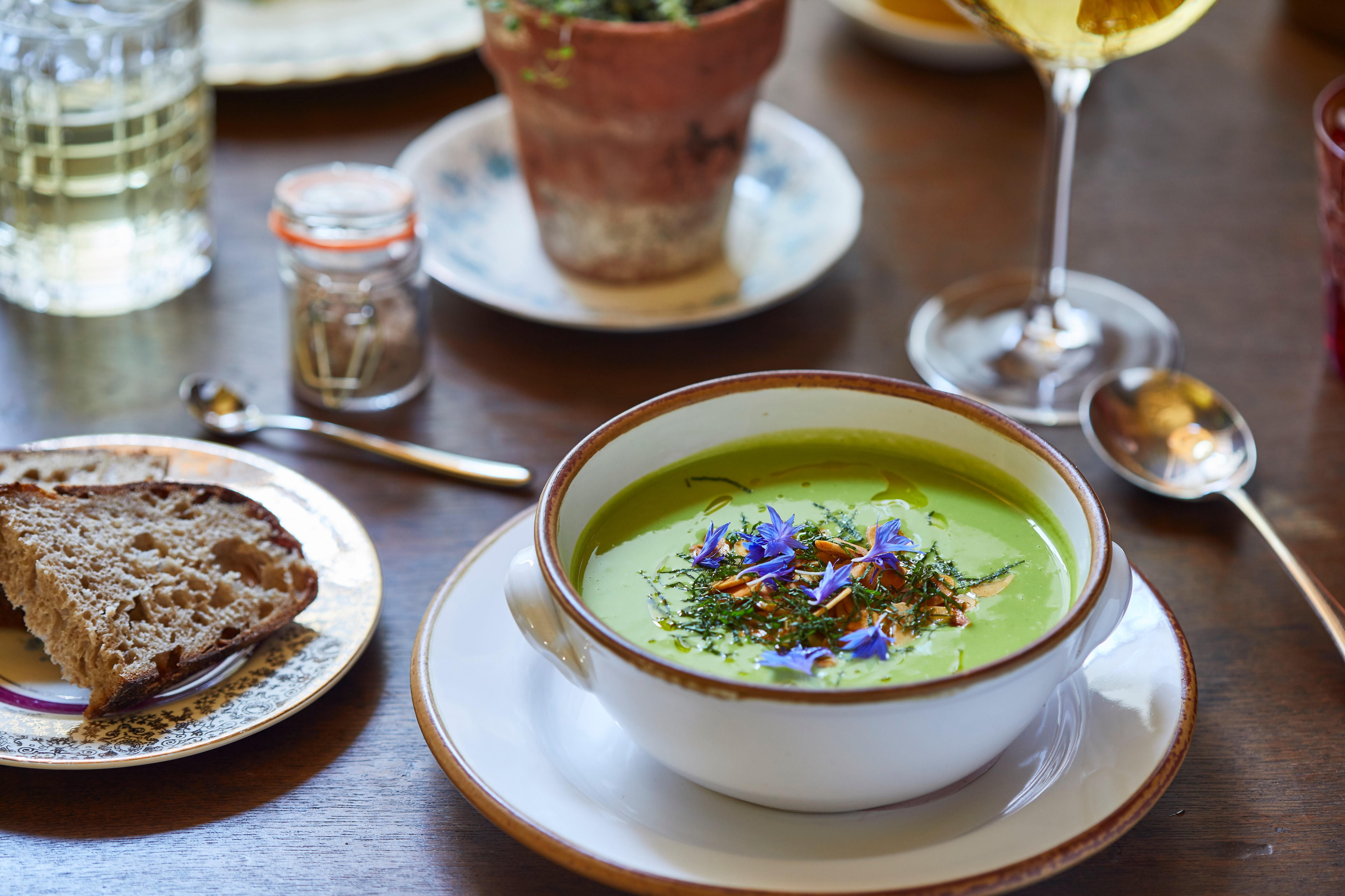 Pea soup garnished with flowers on a table with bread, a spoon and a glass of wine. A flower pot stands in the background.