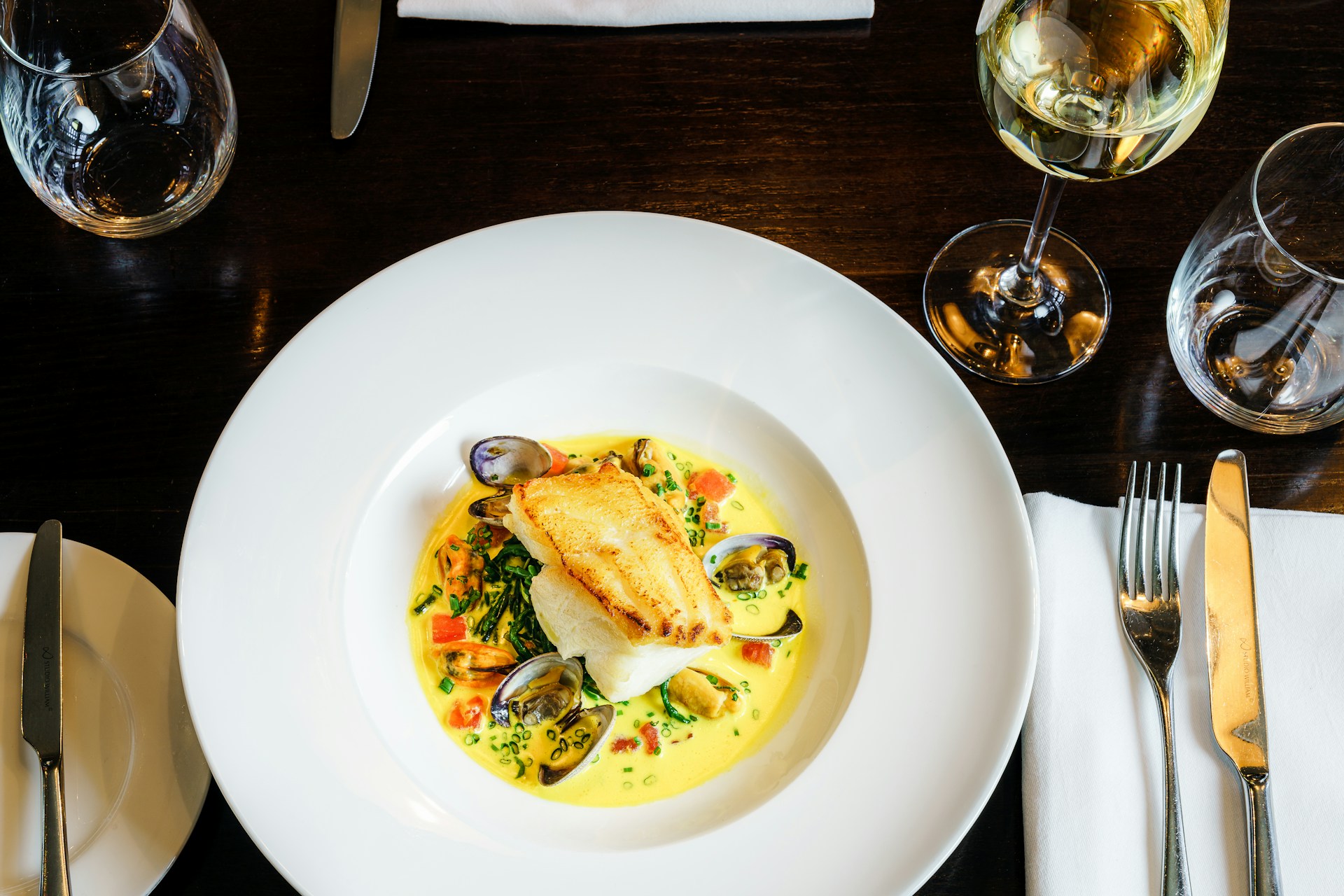 Overhead shot of wide-rimmed white bowl on wooden table top, with pan-fried white fish and mussels in a creamy sauce, accompanied by glass of white wine, linen napkin and cutlery