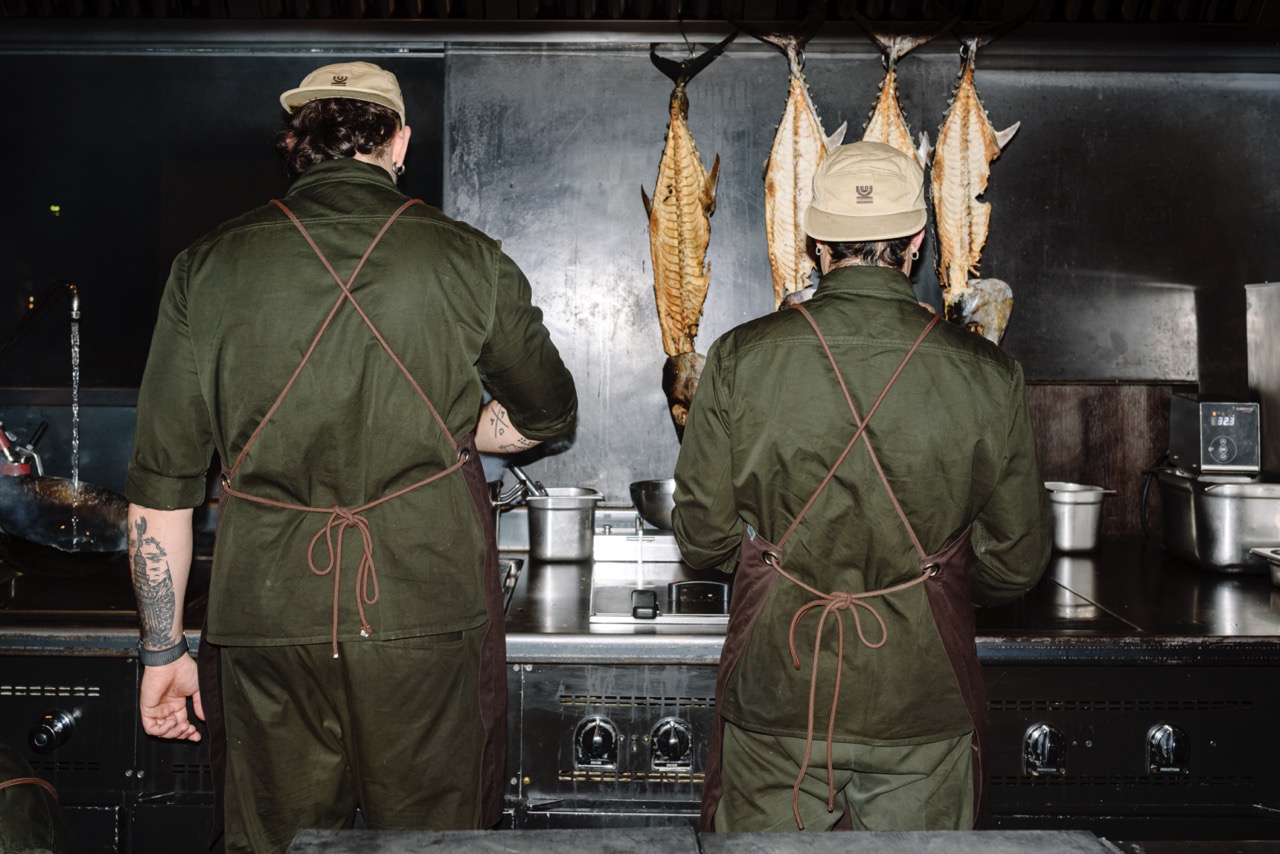The backs of two chefs standing at the stove in dark green uniforms, with fish hanging in front of them
