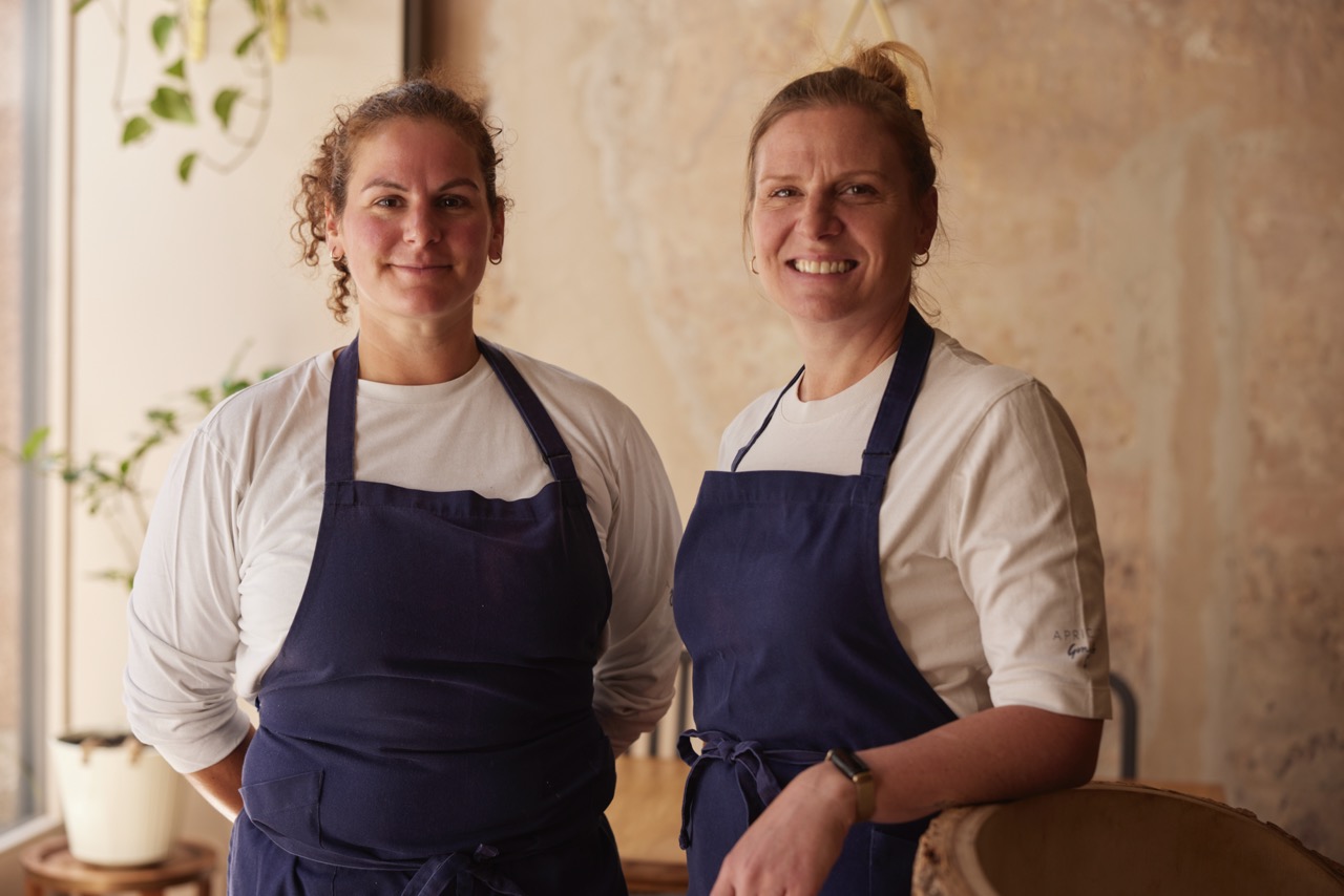 Chefs Eve Seemann & Chantelle Nicholson in Apricity, wearing white chef jackets and navy aprons, smiling for camera