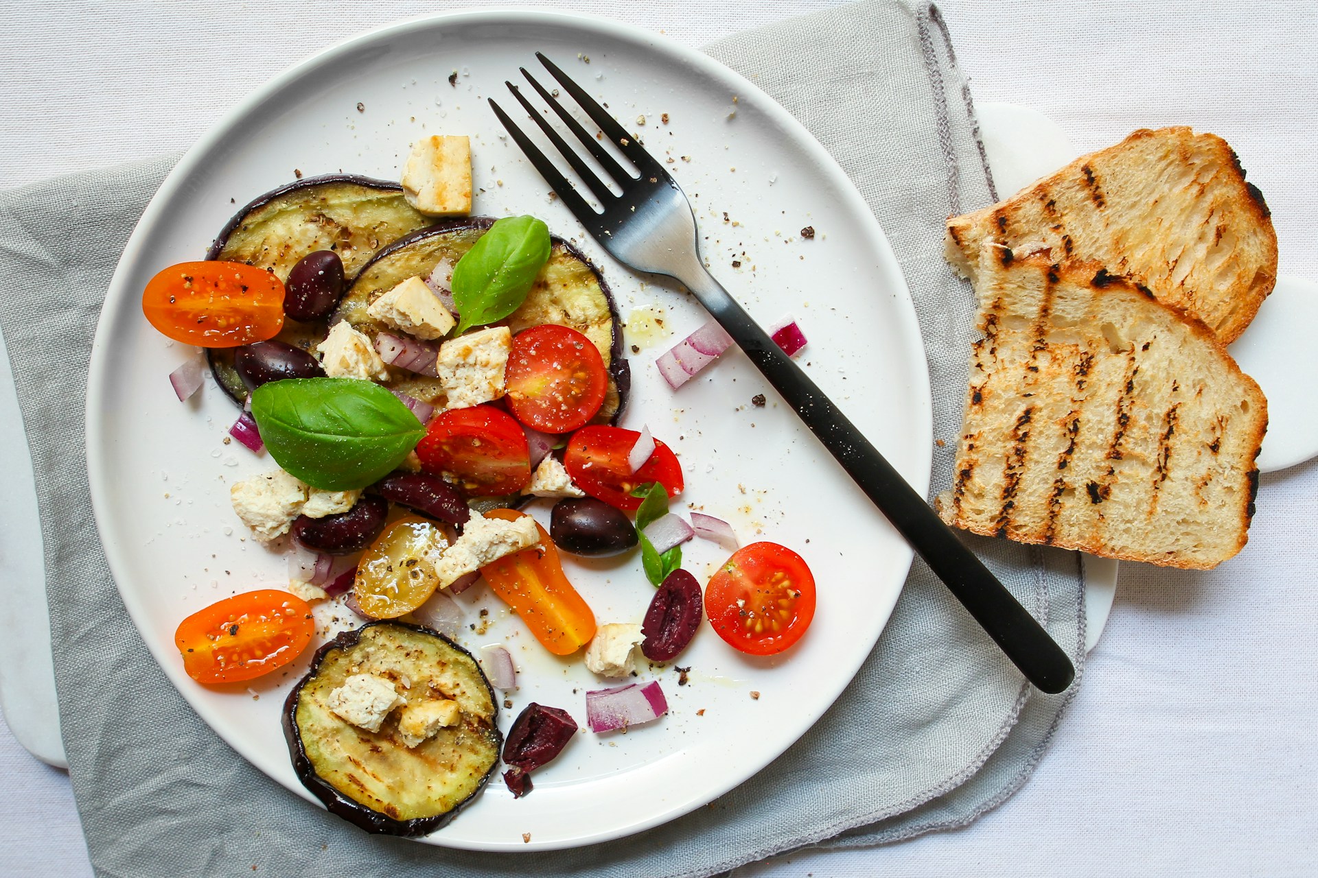 Overhead shot of aubergine, tomatoes, red onion and basil on plain white plate sitting on folded pale blue cloth napkin. Two lightly toasted slices of bread sit beside the plate.