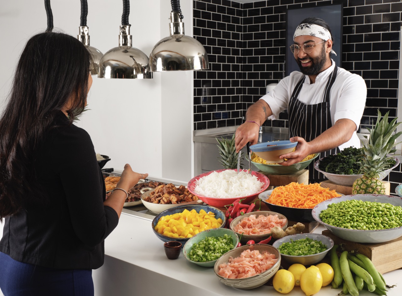 Smiling Restaurant Associates staff member in chef jacket and apron serving customer from colourful salad bar