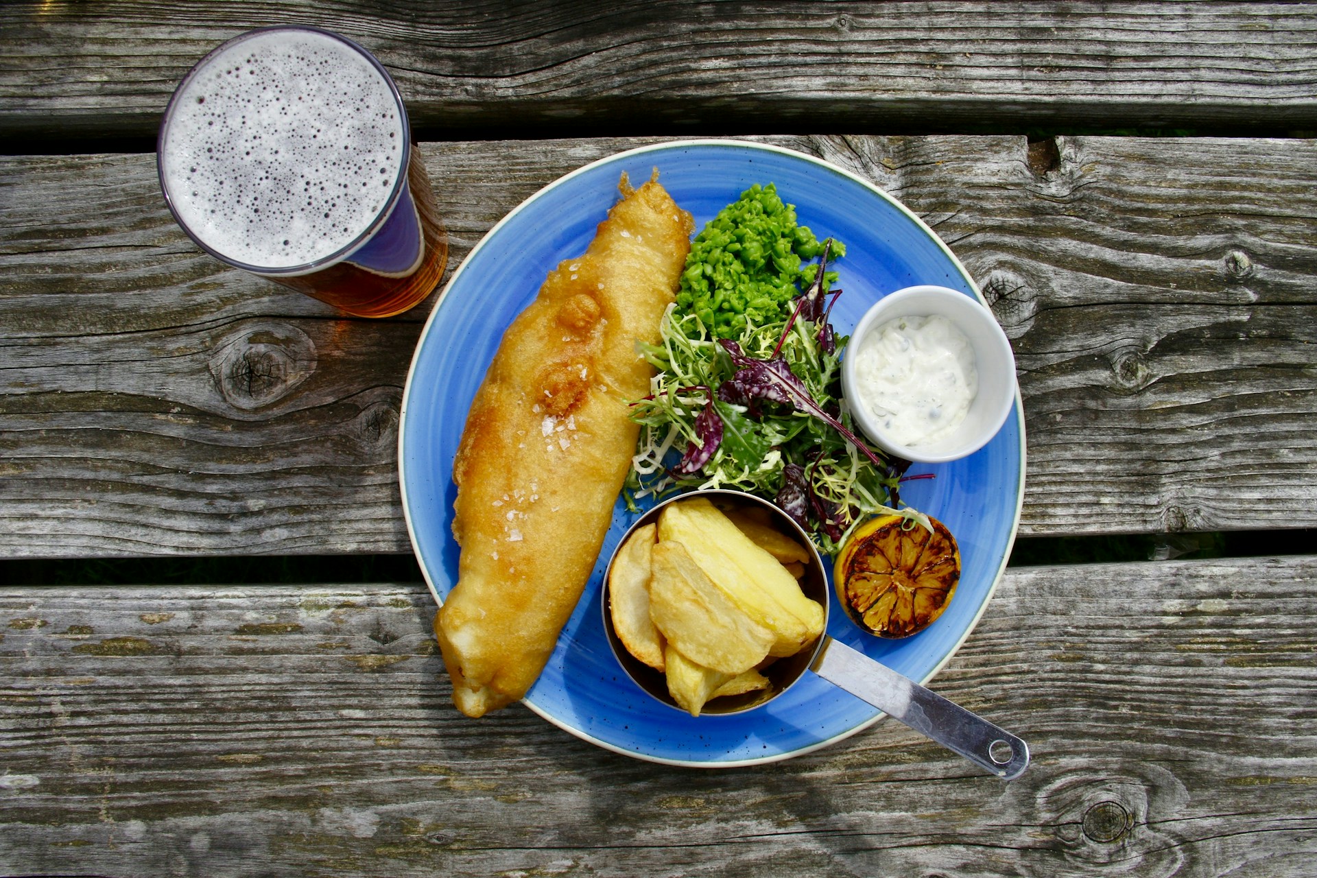 Overhead shot of a blue plate with battered fish, chips, salad, peas, tartare sauce and a lemon, with a glass of beer, all on a wooden table