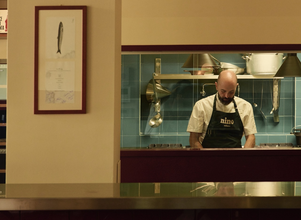 A chef in an open kitchen viewed from the restaurant floor, wearing an apron that says "Nino"