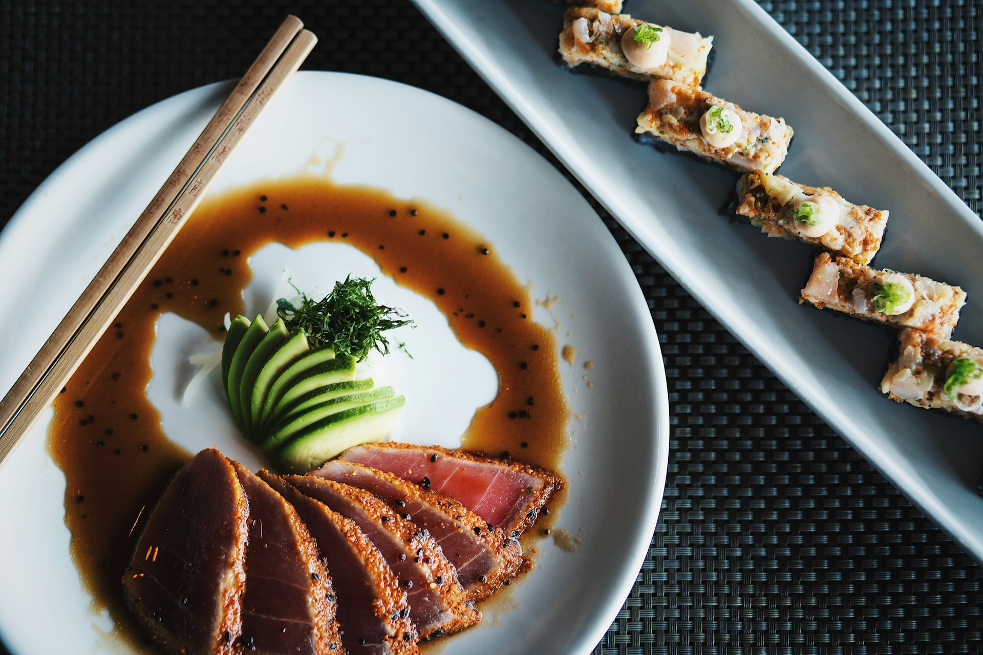 Overhead shot of sliced raw tuna with soy sauce and sesame seeds, with sushi on separate dish