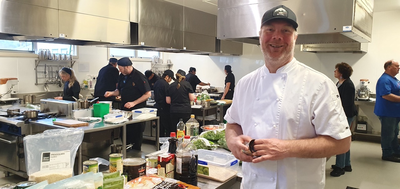 A chef smiling in a University Of Otago kitchen with more chefs busy in the background