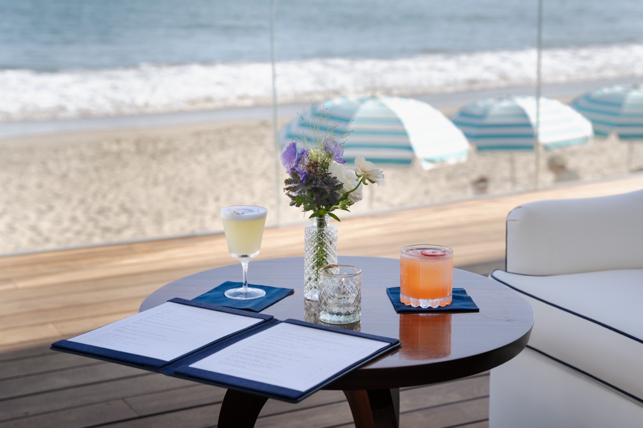 An outdoor table at Caruso's Rosewood Miramar Beach, with menu and cocktails, with beach umbrellas in the background