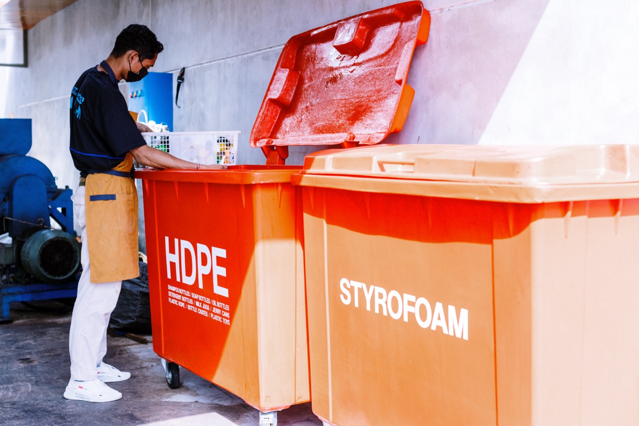 A worker sorting waste into two big orange bins, one saying HDPE and one saying Styrofoam