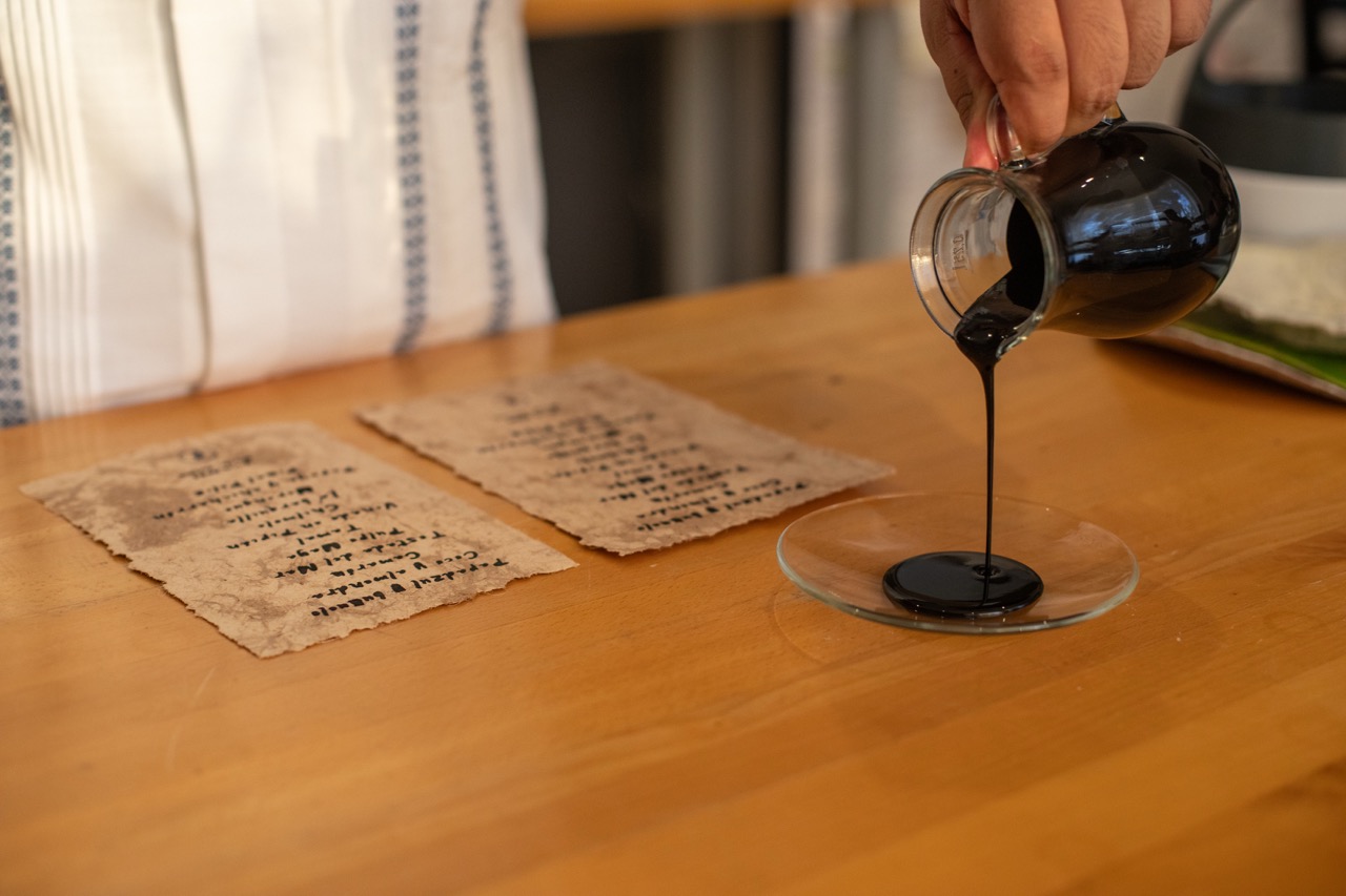 A hand pouring black liquid from a jug into a small glass dish at K'U'UK