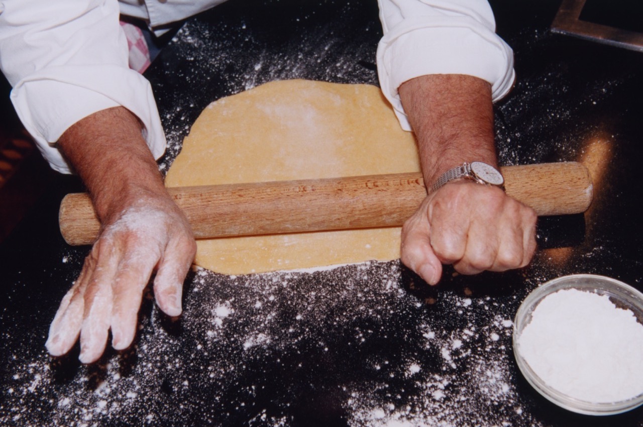 Hands using a rolling pin to roll out pastry on dark, floured surface