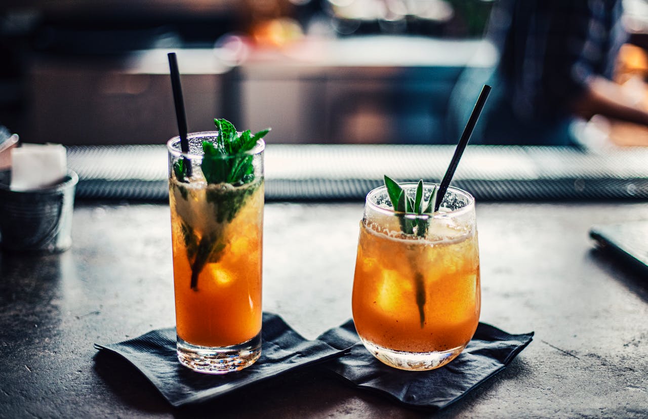 Two bright cocktails garnished with rosemary, served on black napkins on dark bar counter