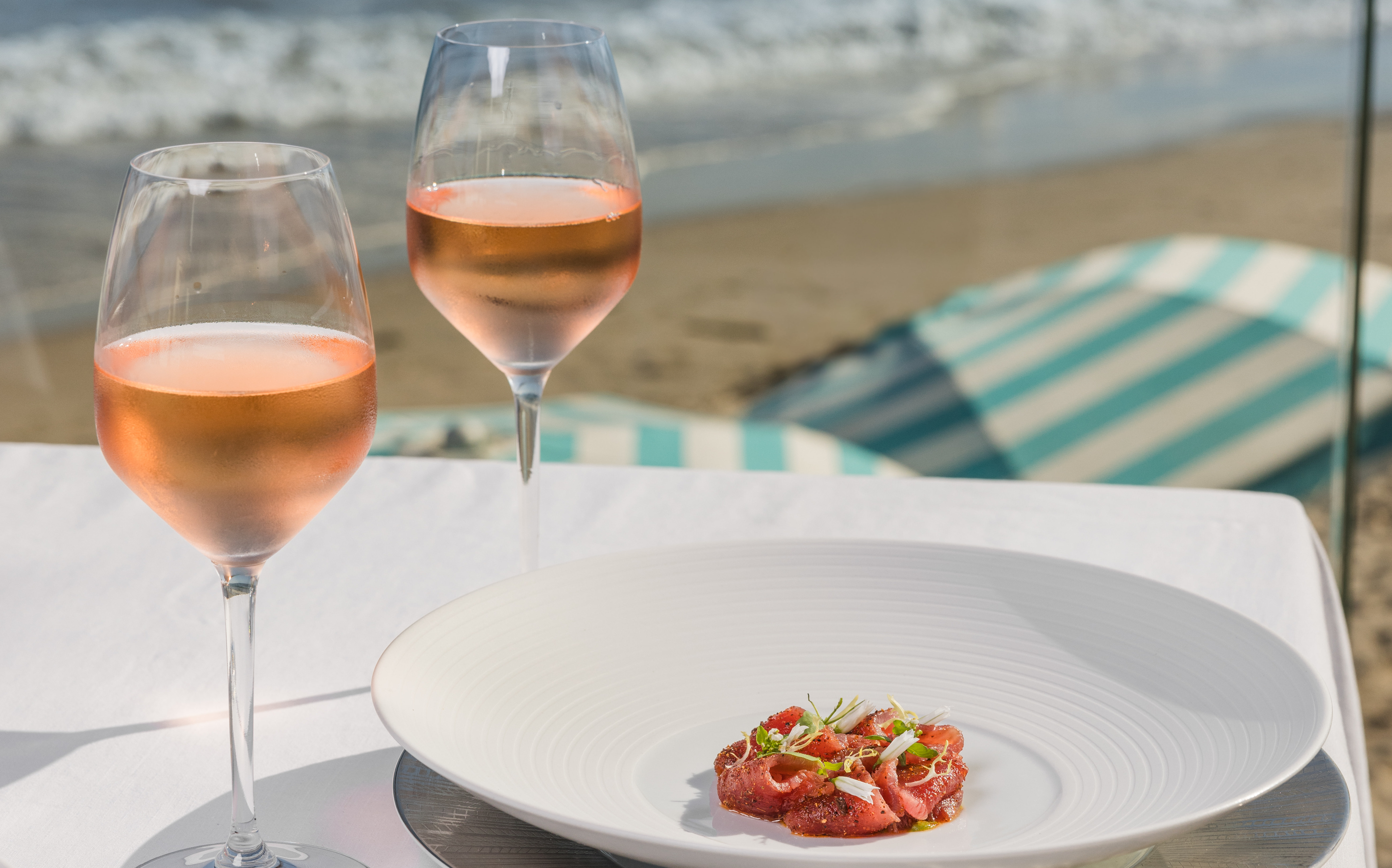 Seafood dish in shallow white bowl on table with two glasses of rose wine. Beach and sea in background.