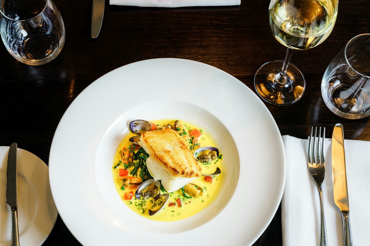 Overhead shot of fish dish with clams on a wooden table with a glass of white wine