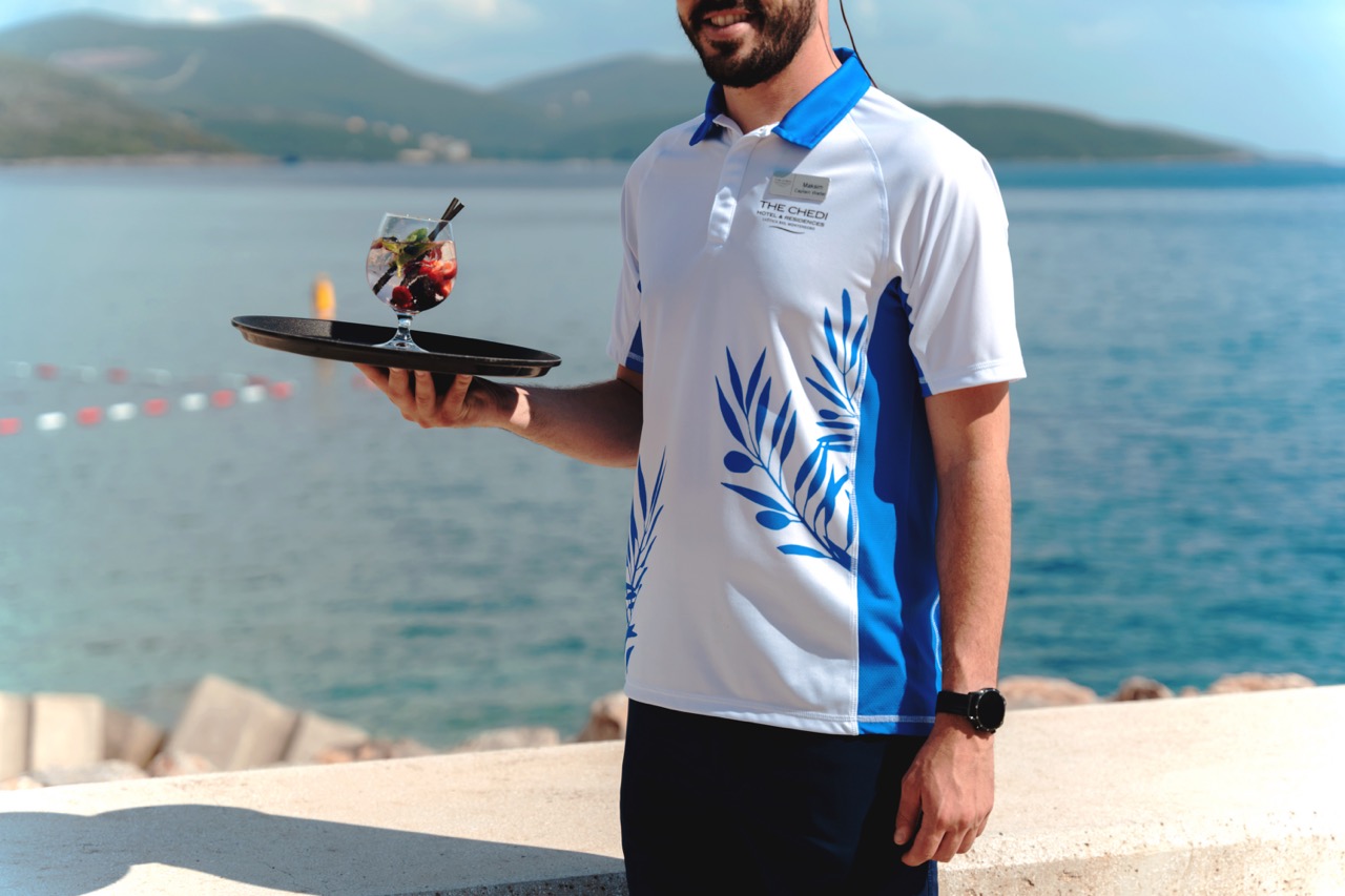 A smiling waiter outdoors in sunshine, wearing a blue and white uniform top, carrying a cocktail on a tray 