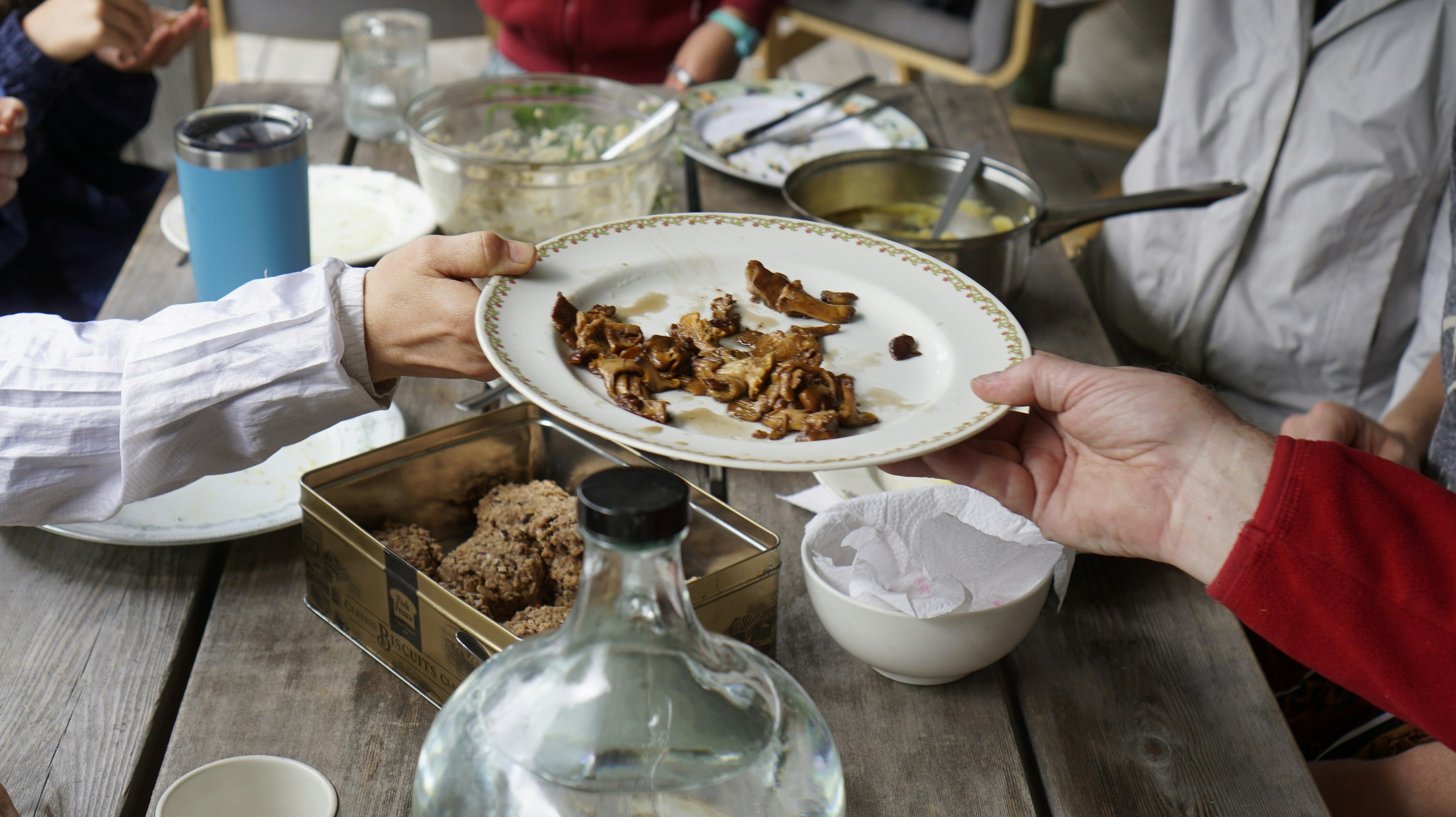 People sharing food at a wooden table, with two people passing a plate of mushrooms between them