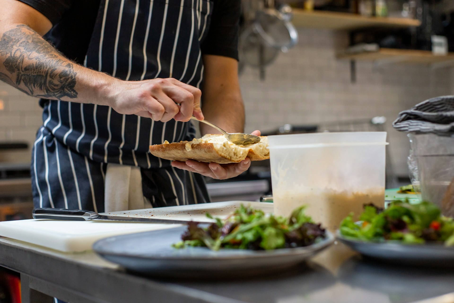 A chef in a striped apron preparing food, spooning something into a baguette with salads in foreground