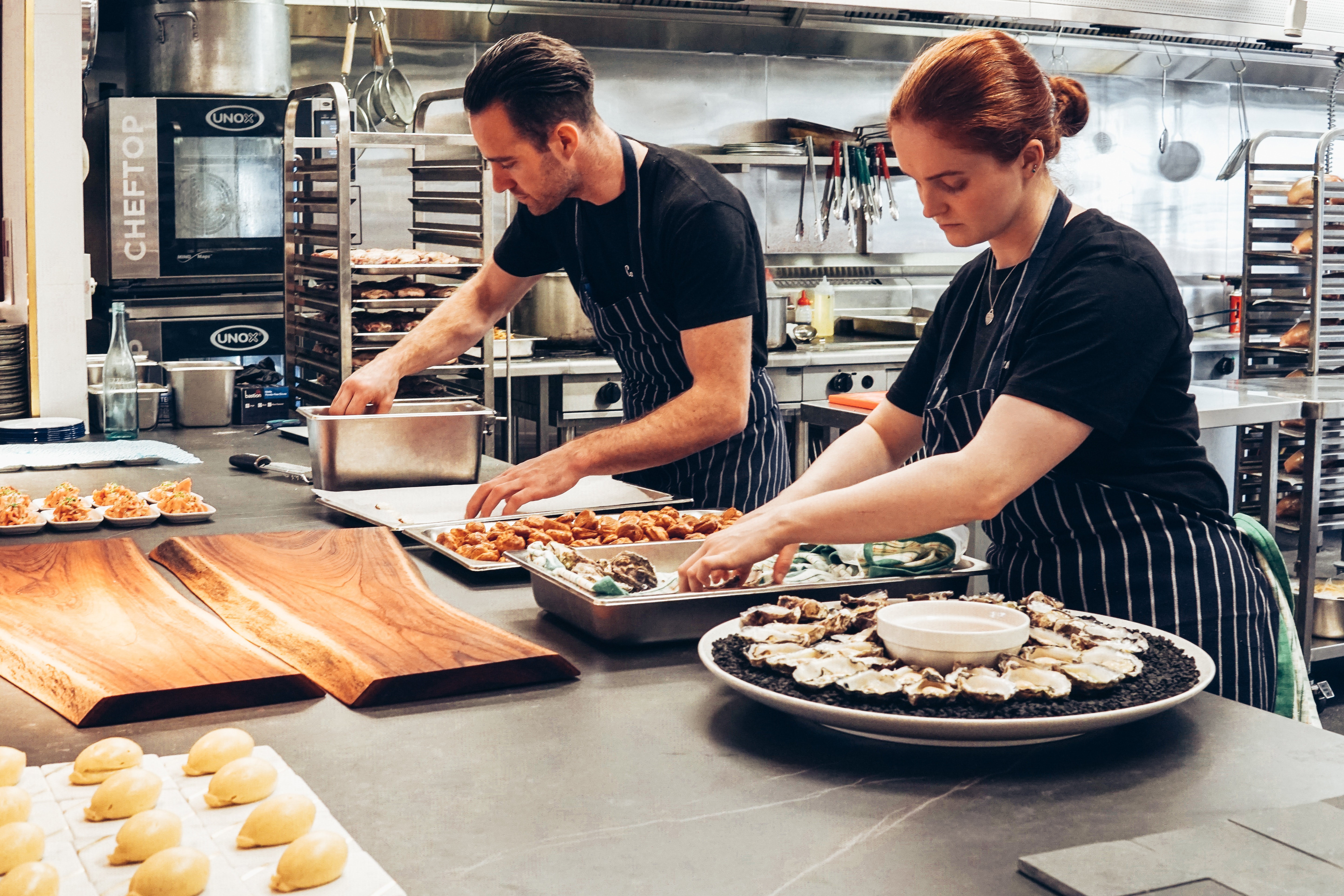 Two chefs in black t-shirts and striped aprons preparing food in the kitchen