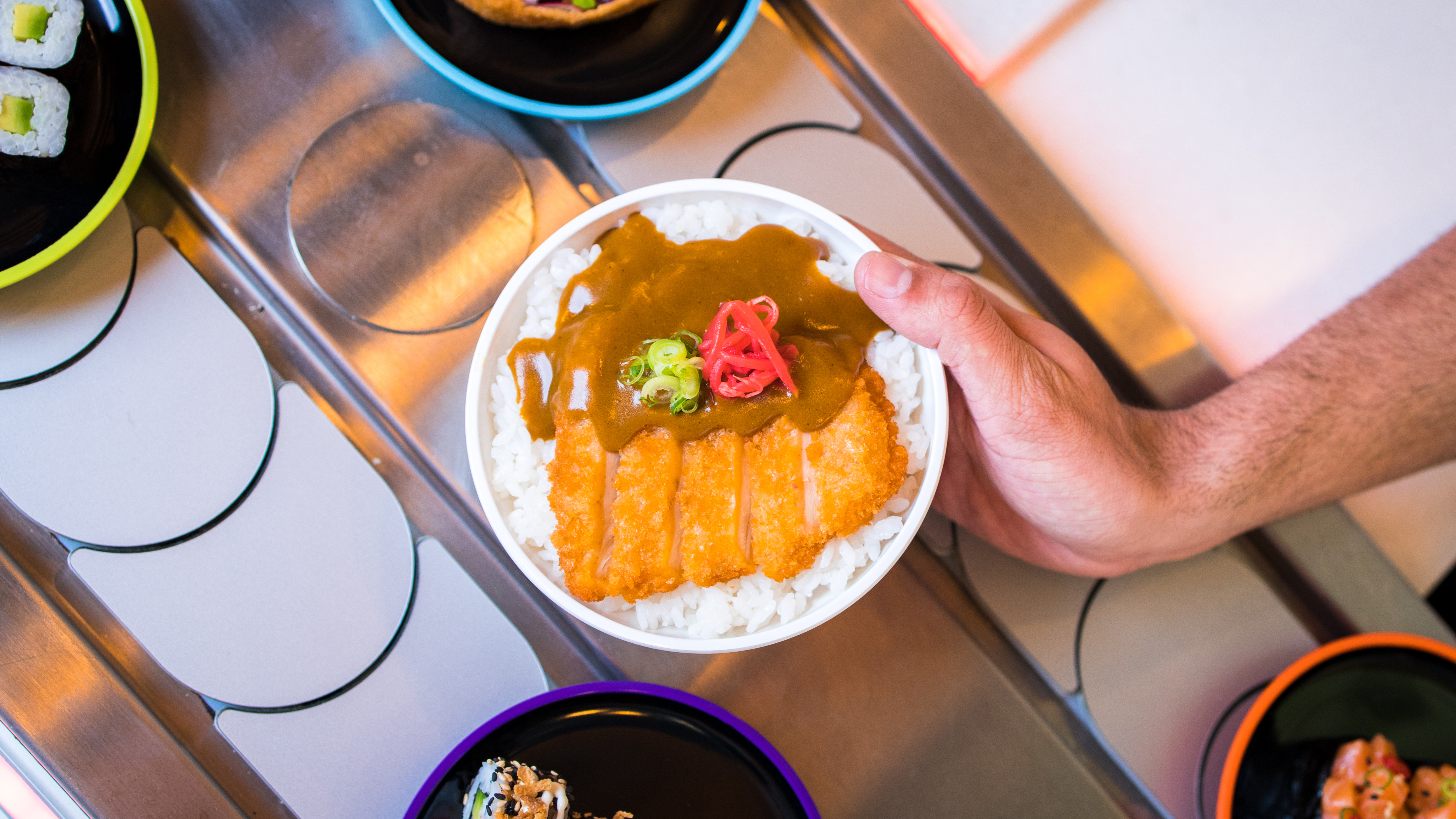 Hand holding plate of katsu curry over conveyor belt at Yo! Sushi