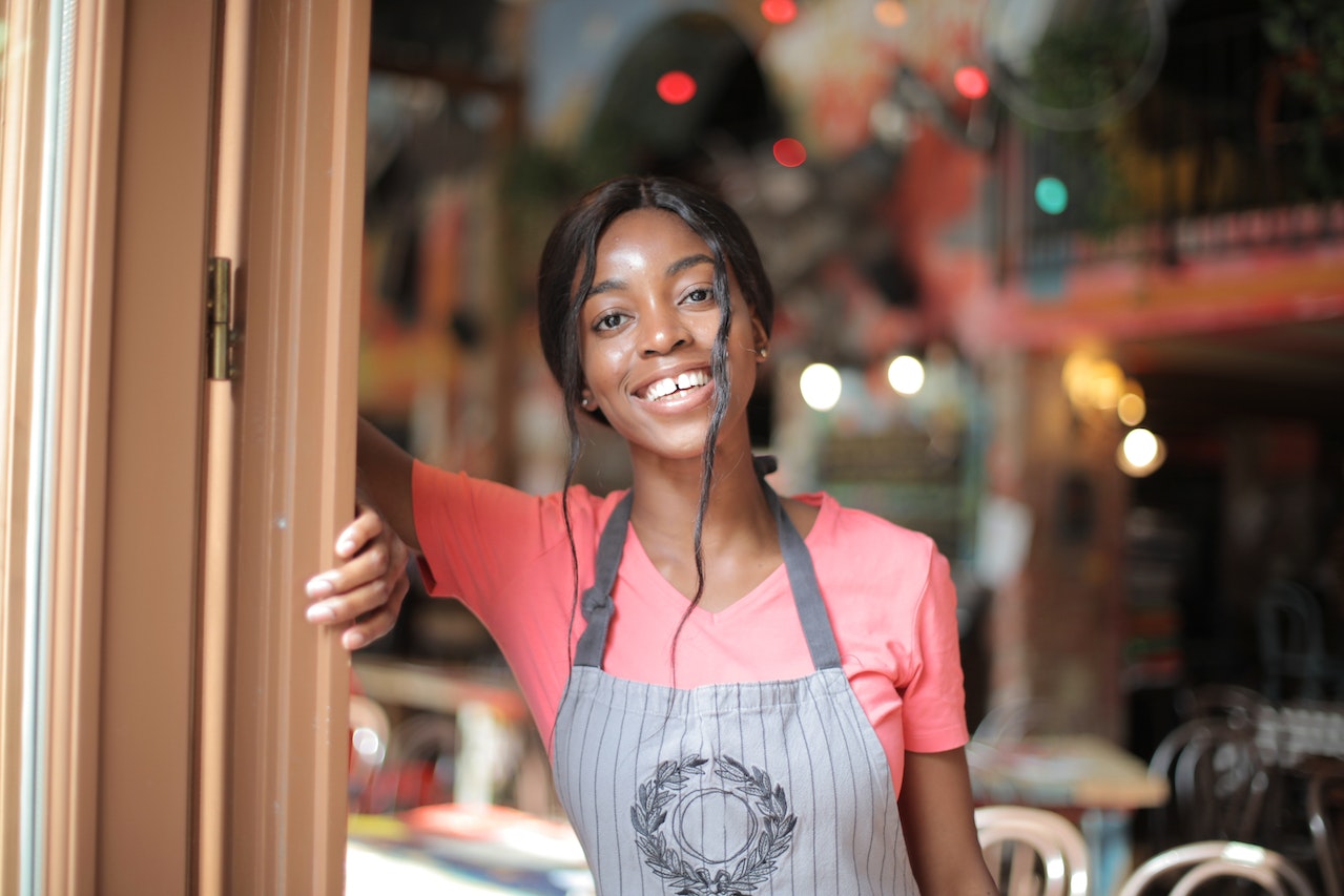 Happy waitress wearing pink t shirt and grey apron, smiling at camera with restaurant behind her